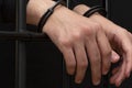 The hands of a young man in handcuffs hang over the bars. Dark tone.