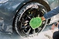 Hands of young man in jeans shirt holding green sponge, washing car wheel with foam. Cleaning of modern rims of luxury Royalty Free Stock Photo
