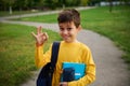 Handsome adorable schoolboy showing singn OK with his hand, standing with backpack and school supplies in the city park