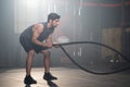 Handsome adult Caucasian men swing battler ropes to do crossfit exercise inside of fitness gym to workout for body strength and