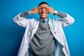 Handsome african american doctor man wearing coat and stethoscope over blue background covering eyes with hands smiling cheerful Royalty Free Stock Photo