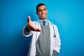 Handsome african american doctor man wearing coat and stethoscope over blue background smiling cheerful offering palm hand giving Royalty Free Stock Photo