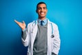 Handsome african american doctor man wearing coat and stethoscope over blue background smiling cheerful presenting and pointing Royalty Free Stock Photo