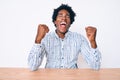 Handsome african american man with afro hair wearing casual clothes sitting on the table very happy and excited doing winner Royalty Free Stock Photo