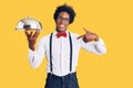 Handsome african american man with afro hair wearing waiter uniform holding silver tray pointing finger to one self smiling happy Royalty Free Stock Photo