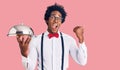 Handsome african american man with afro hair wearing waiter uniform holding silver tray pointing thumb up to the side smiling Royalty Free Stock Photo