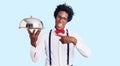 Handsome african american man with afro hair wearing waiter uniform holding silver tray smiling happy pointing with hand and Royalty Free Stock Photo