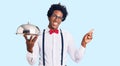 Handsome african american man with afro hair wearing waiter uniform holding silver tray smiling happy pointing with hand and Royalty Free Stock Photo