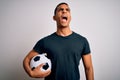 Handsome african american man playing footbal holding soccer ball over white background angry and mad screaming frustrated and Royalty Free Stock Photo