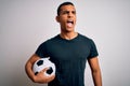 Handsome african american man playing footbal holding soccer ball over white background angry and mad screaming frustrated and Royalty Free Stock Photo