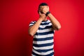 Handsome african american man wearing casual striped t-shirt standing over red background Covering eyes and mouth with hands, Royalty Free Stock Photo