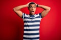 Handsome african american man wearing casual striped t-shirt standing over red background Crazy and scared with hands on head, Royalty Free Stock Photo