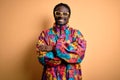 Handsome african american man wearing colorful coat and glasses over yellow background happy face smiling with crossed arms Royalty Free Stock Photo