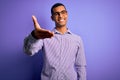 Handsome african american man wearing striped shirt and glasses over purple background smiling friendly offering handshake as