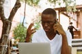 Handsome afro american man with glasses sitting at table in cafe on summer veranda, talking on telephone and working on Royalty Free Stock Photo