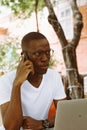 Handsome afro american man with glasses sitting at table in cafe on summer veranda, talking on telephone and working on Royalty Free Stock Photo