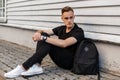 Handsome American man with a stylish hairstyle in a fashionable black T-shirt in stylish jeans in white sneakers with a backpack Royalty Free Stock Photo