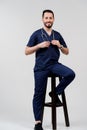 Handsome arab doctor surgeon smiles on white background. Bearded student with stethoscope in medical robe seats on chair