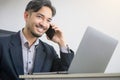 Handsome Asian business man in a suit is sitting and talking on his mobile phone at his desk with a laptop computer lying on it, Royalty Free Stock Photo