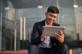Handsome Asian businessman using his tablet while sitting on stairs outside of the company building Royalty Free Stock Photo