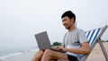 A handsome Asian man sits in a chair on his laptop computer by the sea as the waves crash against the shore, comfortably touching Royalty Free Stock Photo