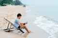 A handsome Asian man sits in a chair on his laptop computer by the sea as the waves crash against the shore, comfortably touching Royalty Free Stock Photo