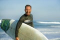 Handsome and attractive surfer man in neoprene swimsuit carrying surf board smiling happy and cheerful after surfing enjoying