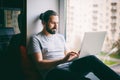Handsome attractive young dark-haired man working on laptop at home while sitting on windowsill with city view Royalty Free Stock Photo