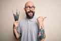 Handsome bald artist man with beard and tattoo painting using painter brushes pointing and showing with thumb up to the side with Royalty Free Stock Photo