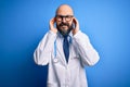 Handsome bald doctor man with beard wearing glasses and stethoscope over blue background covering ears with fingers with annoyed