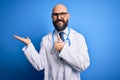 Handsome bald doctor man with beard wearing glasses and stethoscope over blue background Showing palm hand and doing ok gesture Royalty Free Stock Photo