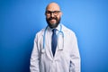 Handsome bald doctor man with beard wearing glasses and stethoscope over blue background winking looking at the camera with sexy Royalty Free Stock Photo