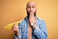 Handsome bald man with beard holding paper airplane over isolated yellow background cover mouth with hand shocked with shame for