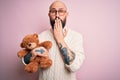 Handsome bald man with beard and tattoo holding teddy bear over isolated pink background cover mouth with hand shocked with shame Royalty Free Stock Photo