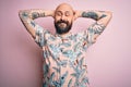 Handsome bald man with beard and tattoo wearing casual floral shirt over pink background relaxing and stretching, arms and hands