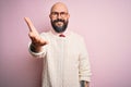 Handsome bald man with beard and tattoo wearing glasses and sweater over pink background smiling friendly offering handshake as