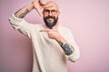 Handsome bald man with beard and tattoo wearing glasses and sweater over pink background smiling making frame with hands and Royalty Free Stock Photo