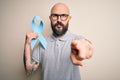 Handsome bald man with beard and tattoos holding blue cancer ribbon over background pointing with finger to the camera