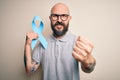 Handsome bald man with beard and tattoos holding blue cancer ribbon over isolated background annoyed and frustrated shouting with