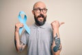 Handsome bald man with beard and tattoos holding blue cancer ribbon over isolated background pointing and showing with thumb up to