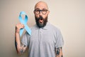 Handsome bald man with beard and tattoos holding blue cancer ribbon over isolated background scared in shock with a surprise face,