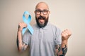 Handsome bald man with beard and tattoos holding blue cancer ribbon over isolated background screaming proud and celebrating