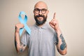 Handsome bald man with beard and tattoos holding blue cancer ribbon over isolated background surprised with an idea or question