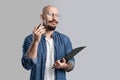 Handsome balded beard with musctache man wearing casual clothes holding clipboard isolated over gray background.