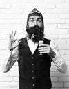 Handsome bearded aviator man with long beard and mustache on funny face holding glass of alcoholic shot in vintage suede Royalty Free Stock Photo