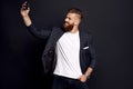 Handsome bearded businessman in suit making selfie isolated on black