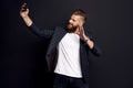 Handsome bearded businessman in suit making selfie isolated on black