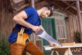 Handsome bearded carpenter in tool belt sawing wooden plank on porch