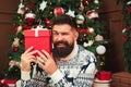 Handsome bearded guy holding christmas gift. Excited happy man in christmas sweater sitting near christmas tree. Christmas paty.