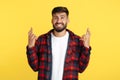 Handsome bearded hipster young man in plaid shirt holding fingers crossed in hope over yellow background. Royalty Free Stock Photo